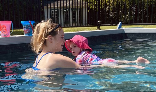 Une monitrice aquatique donne un cours à une petite fille vêtue d'un maillot de bain rose et bleu et d'un chapeau rose, dans une piscine creusée