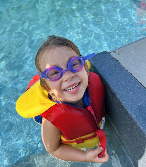 Une élève sourit avec ses lunettes de nageur bleu et rose et son gilet de sauvetage lors d'un cours de natation à domicile